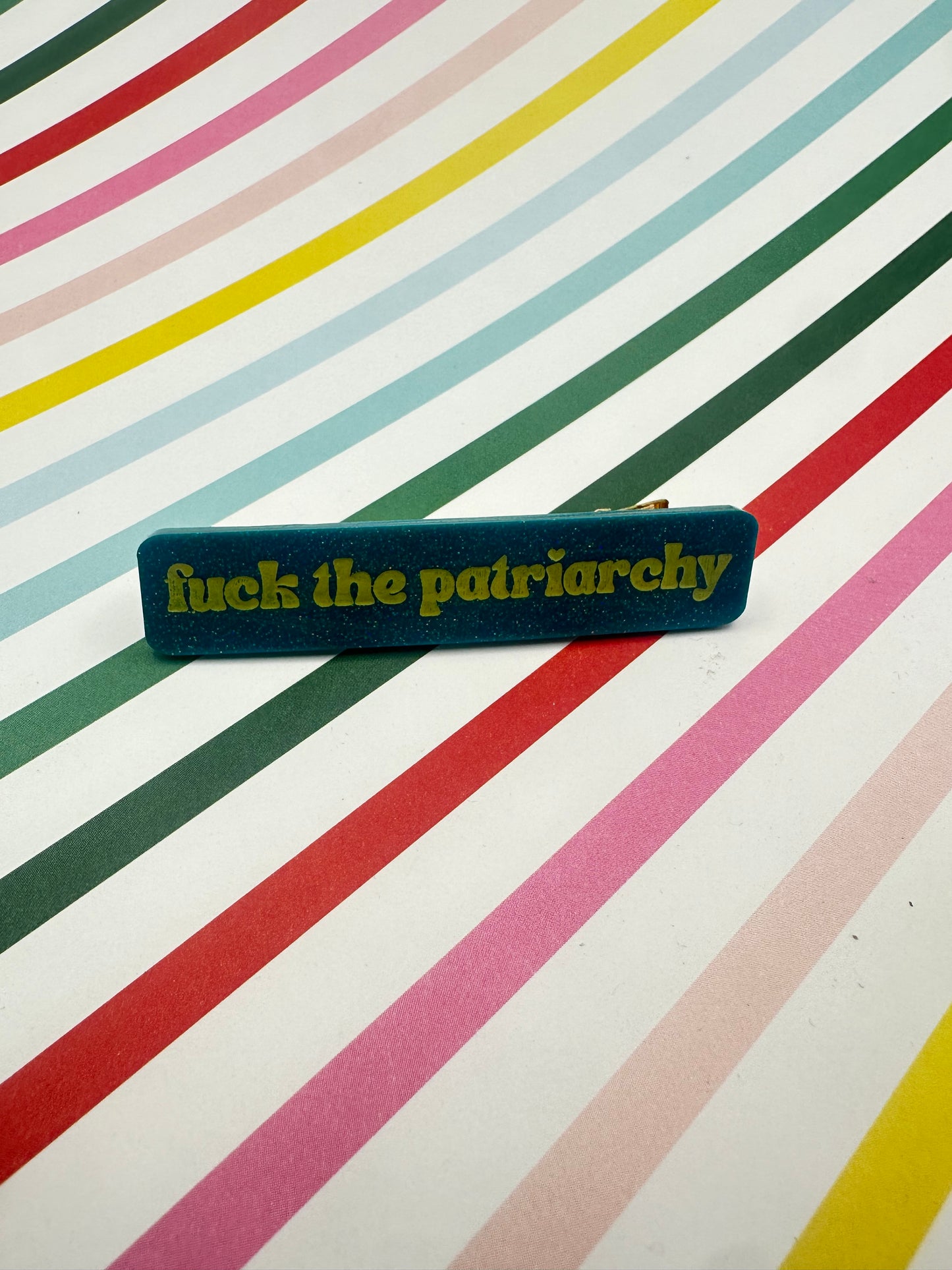 teal hairclip with 'fuck the patriarchy' on a multicolored striped background