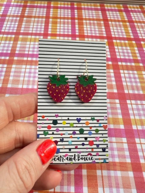 Strawberry-shaped earrings on a striped card held by a hand with red nail polish against a plaid background.