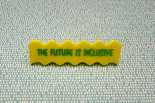 Yellow hairclip with 'The Future is Inclusive' text on a textured fabric background