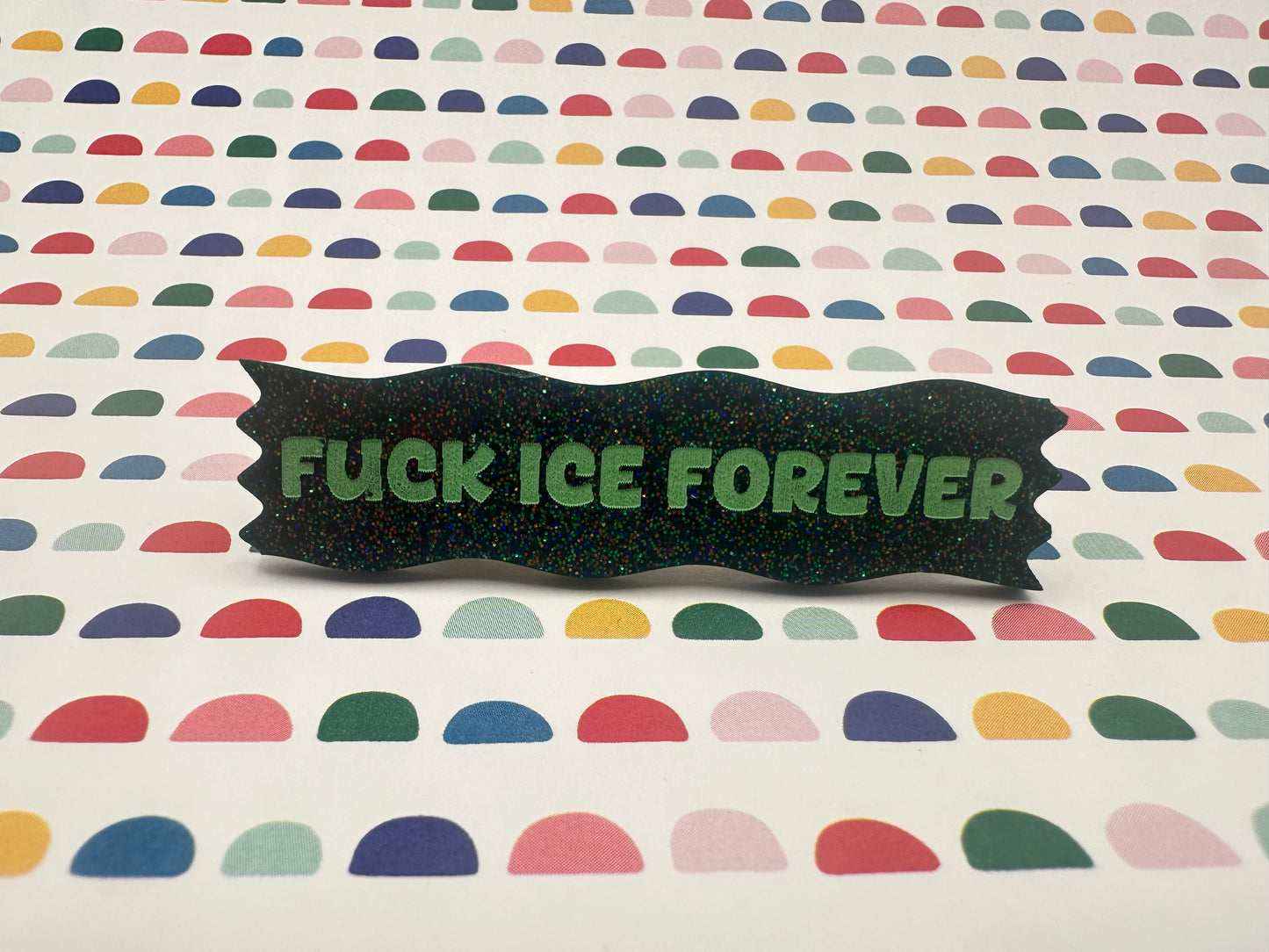 Colorful patterned background with a dark green hairclip displaying 'FUCK ICE FOREVER' in green text.
