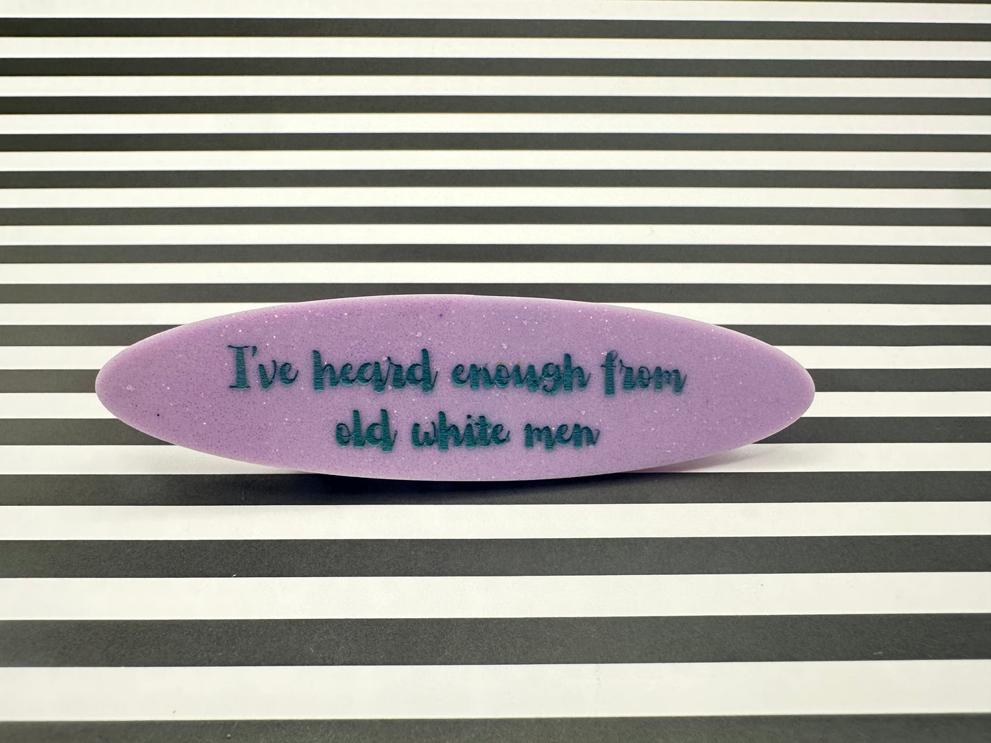 Purple oval hairclip with blue text "I've heard enough from old white men" on a striped background