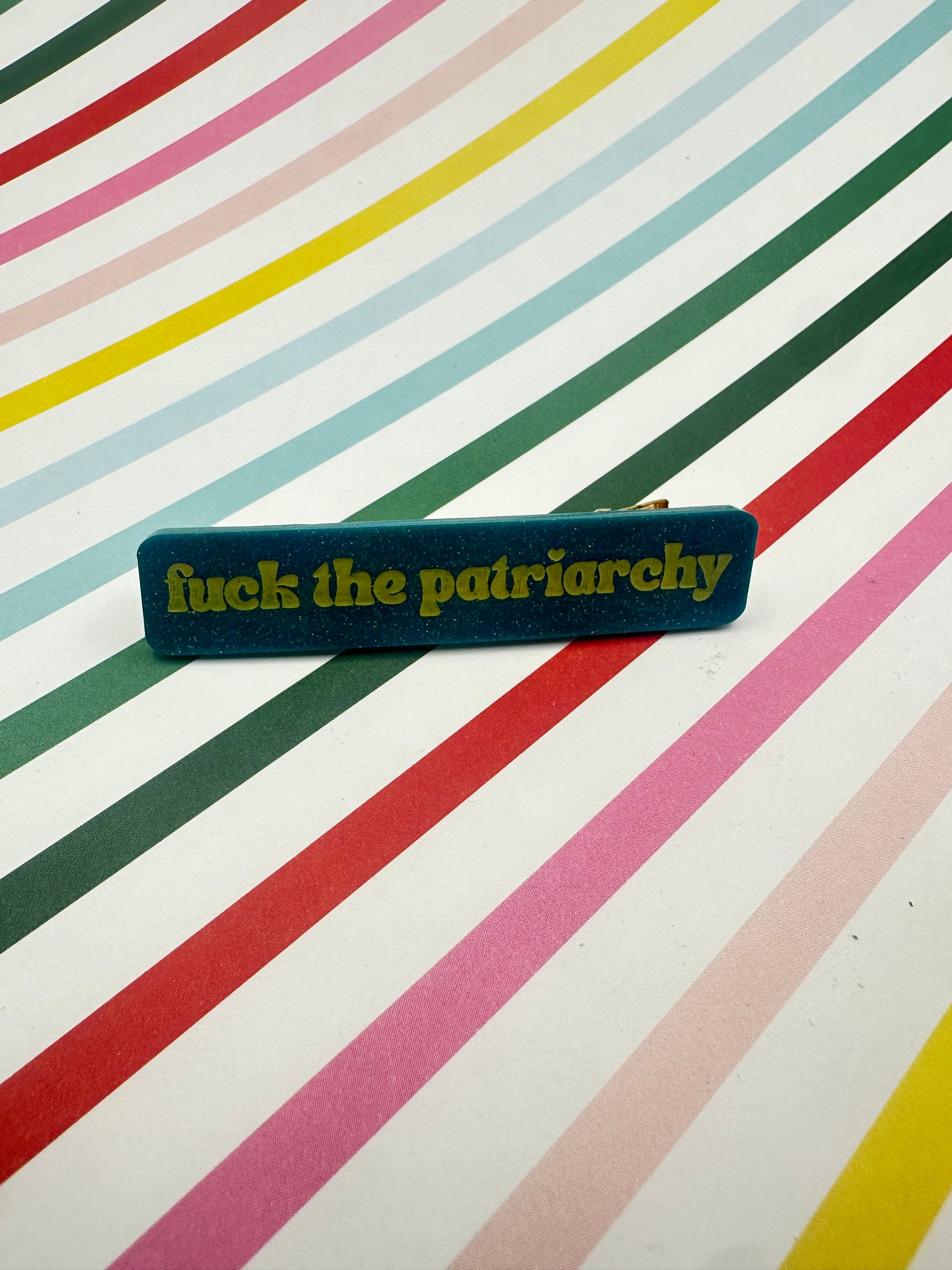 teal hairclip with 'fuck the patriarchy' on a multicolored striped background