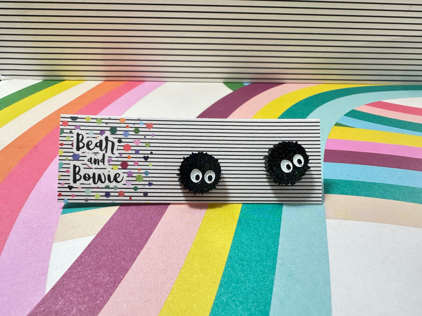Black earrings of anime sprites with eyes on a striped card labeled 'Bear and Bowie' against a colorful striped background.