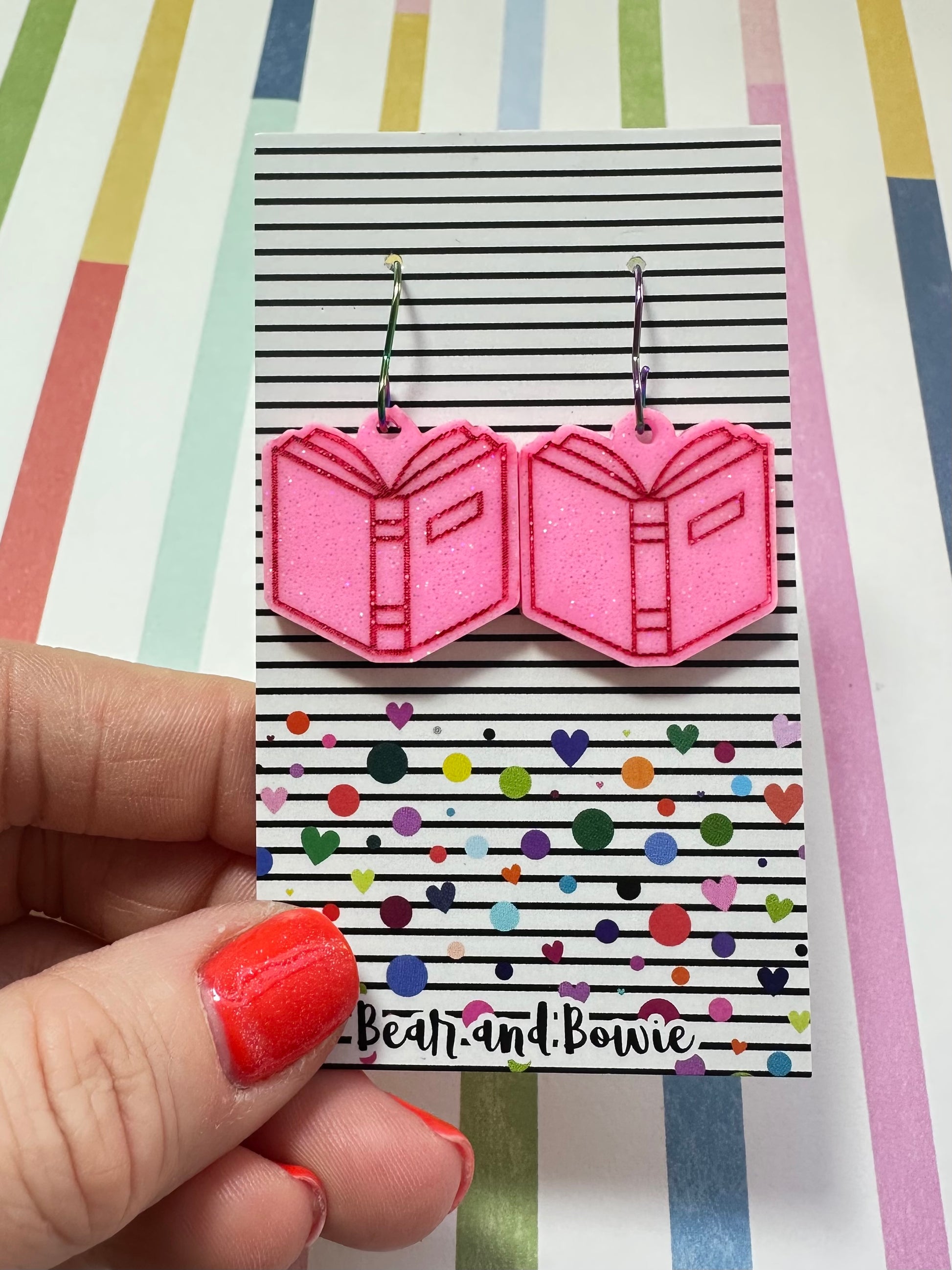 Pink open book earrings on a colorful card held by a hand with red nail polish against a striped background.