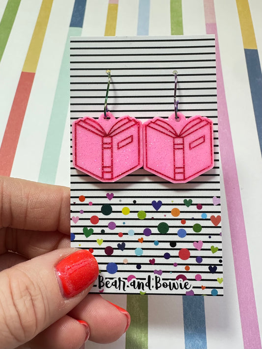 Pink open book earrings on a colorful card held by a hand with red nail polish against a striped background.