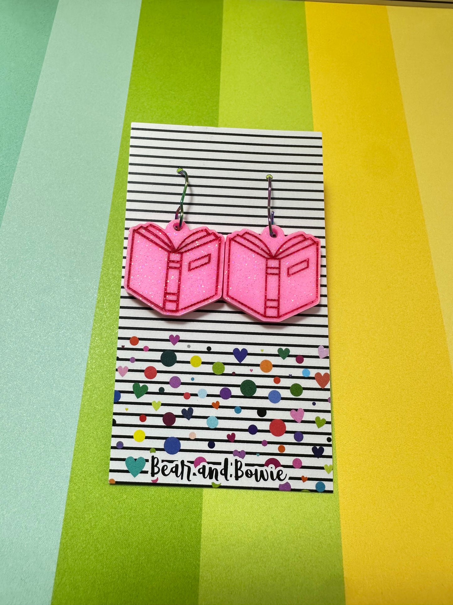 Pink open book earrings on a striped card with colorful dots against a multicolored background