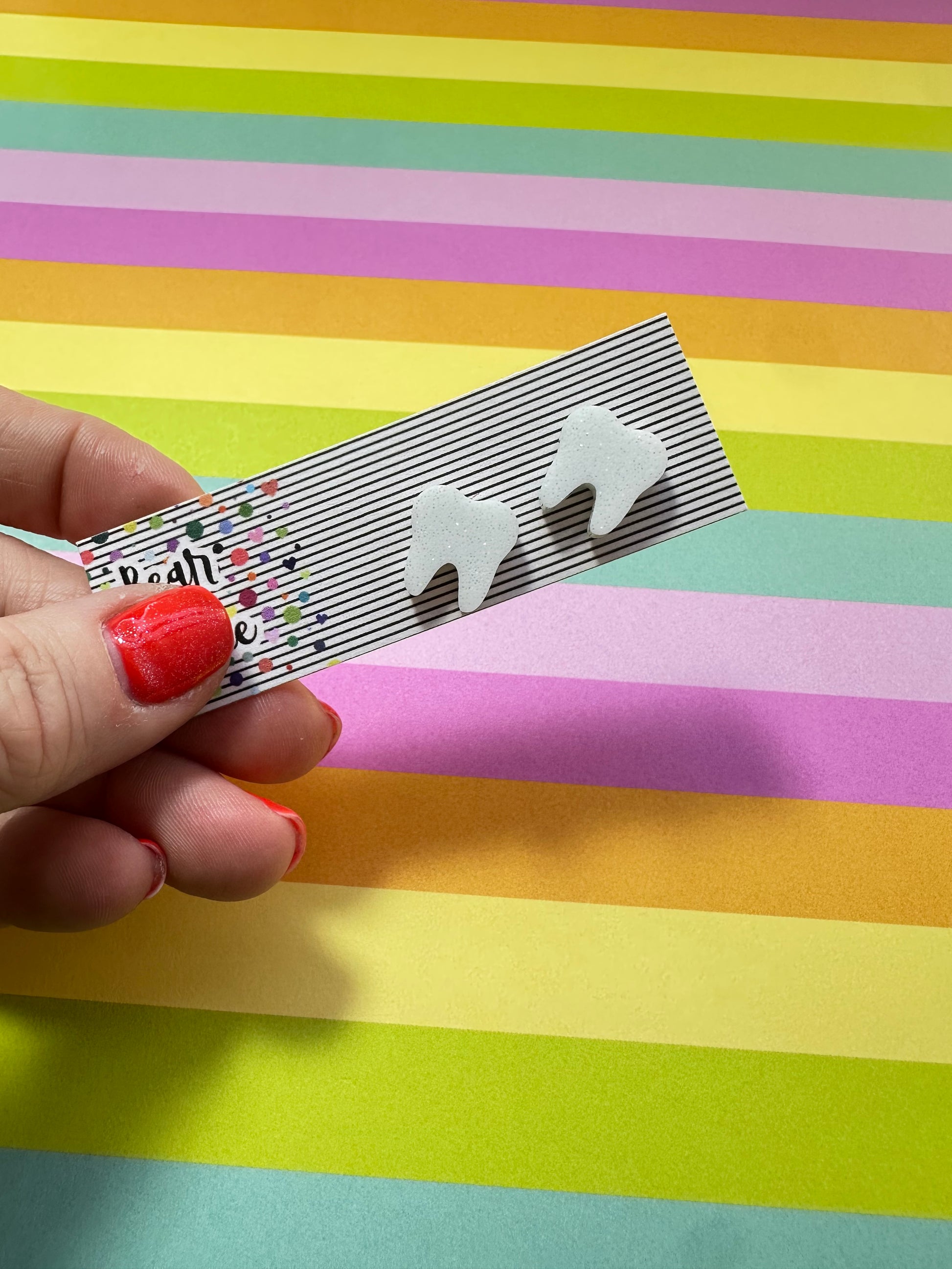 Hand holding a striped product  card with glittery teeth stud earrings against a colorful striped background