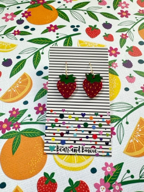 Strawberry-shaped earrings on a striped card with a colorful fruit and floral pattern in the background.