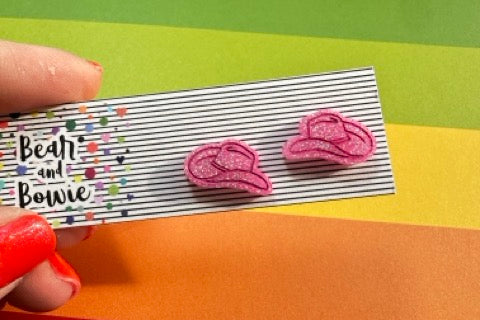 Pink cowgirl hat shaped earrings on a striped card held over a colorful background