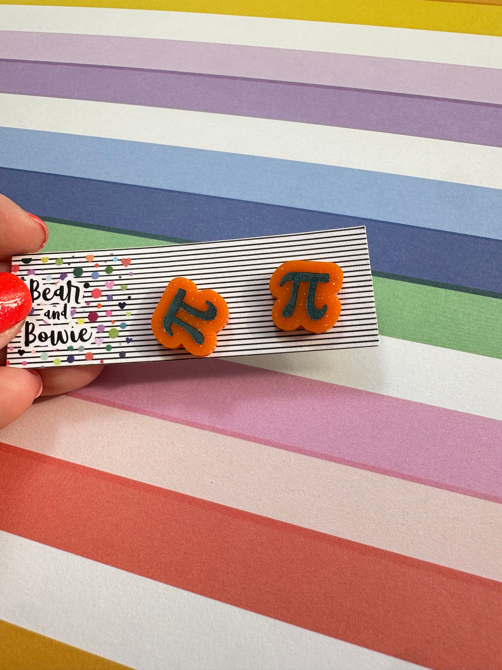 Pair of earrings with etched and painted Pi symbol held over a colorful striped background