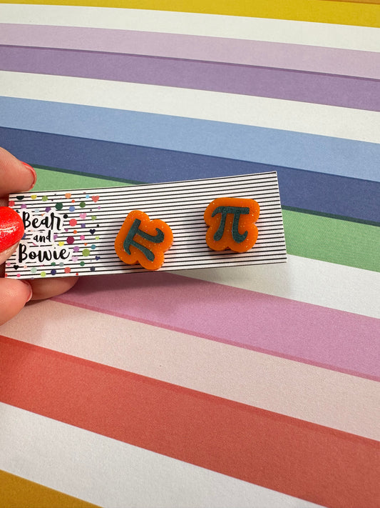 Pair of earrings with etched and painted Pi symbol held over a colorful striped background
