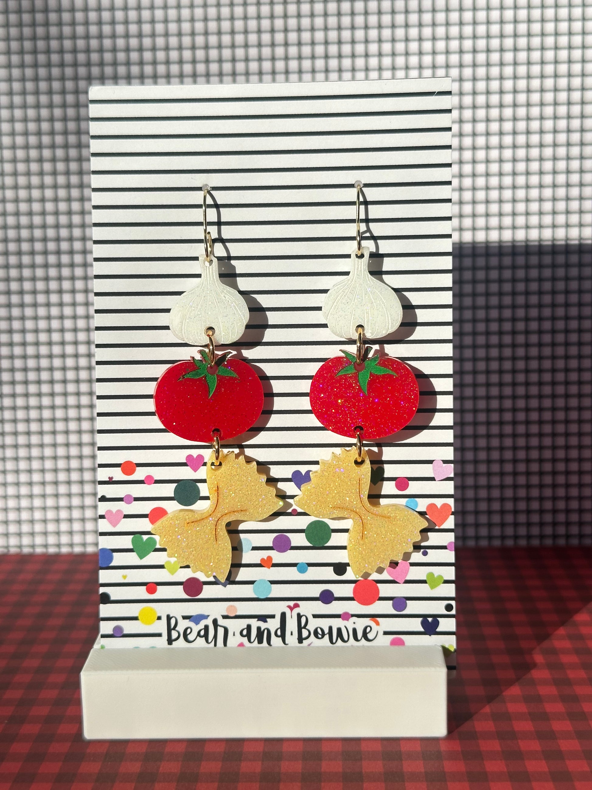 Pair of earrings with white garlic, red tomatoes and pasta on a striped card labeled 'Bear and Bowie'.