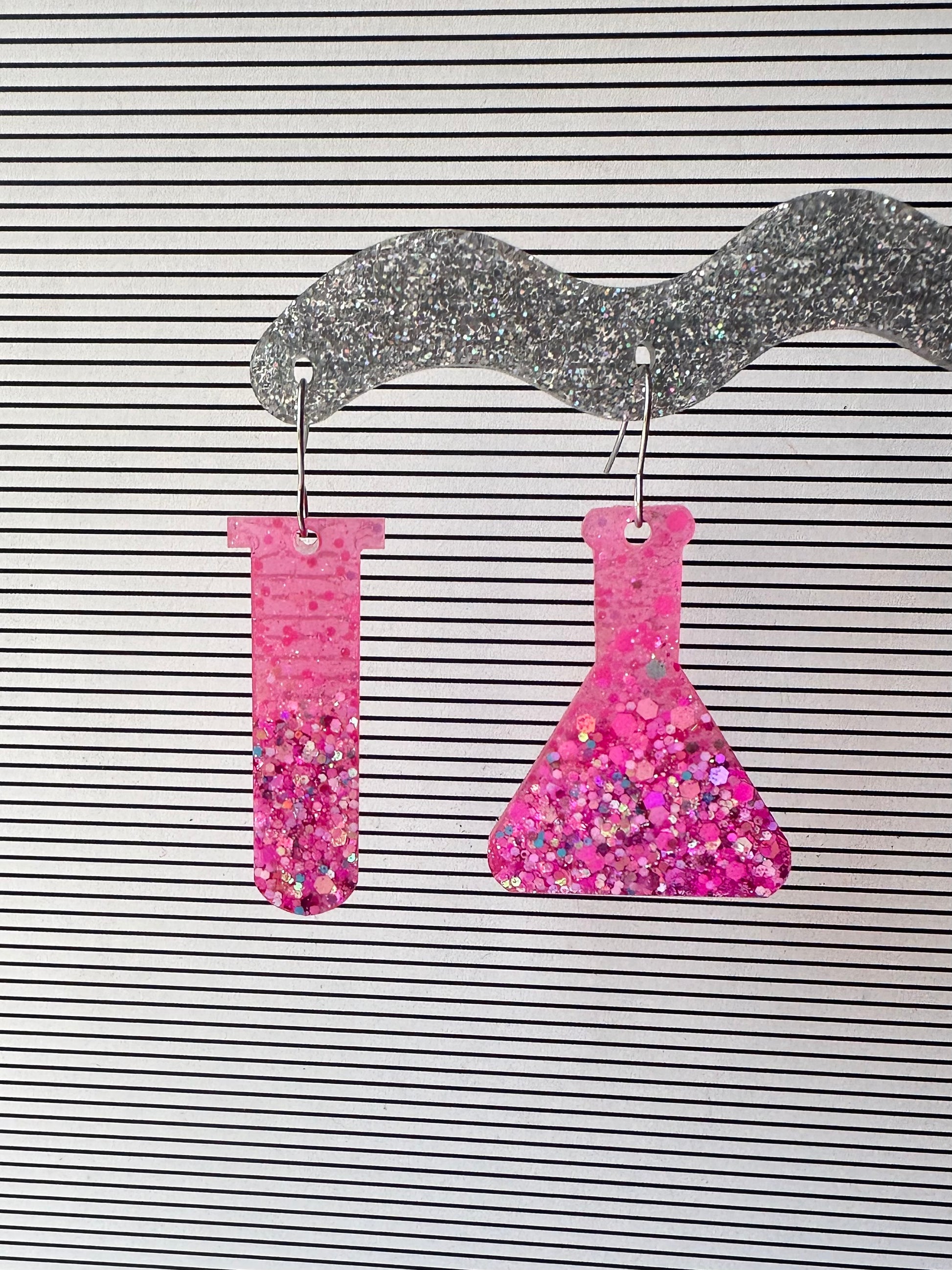 Pink glitter test tube and erlenmeyer flask earrings on a striped background